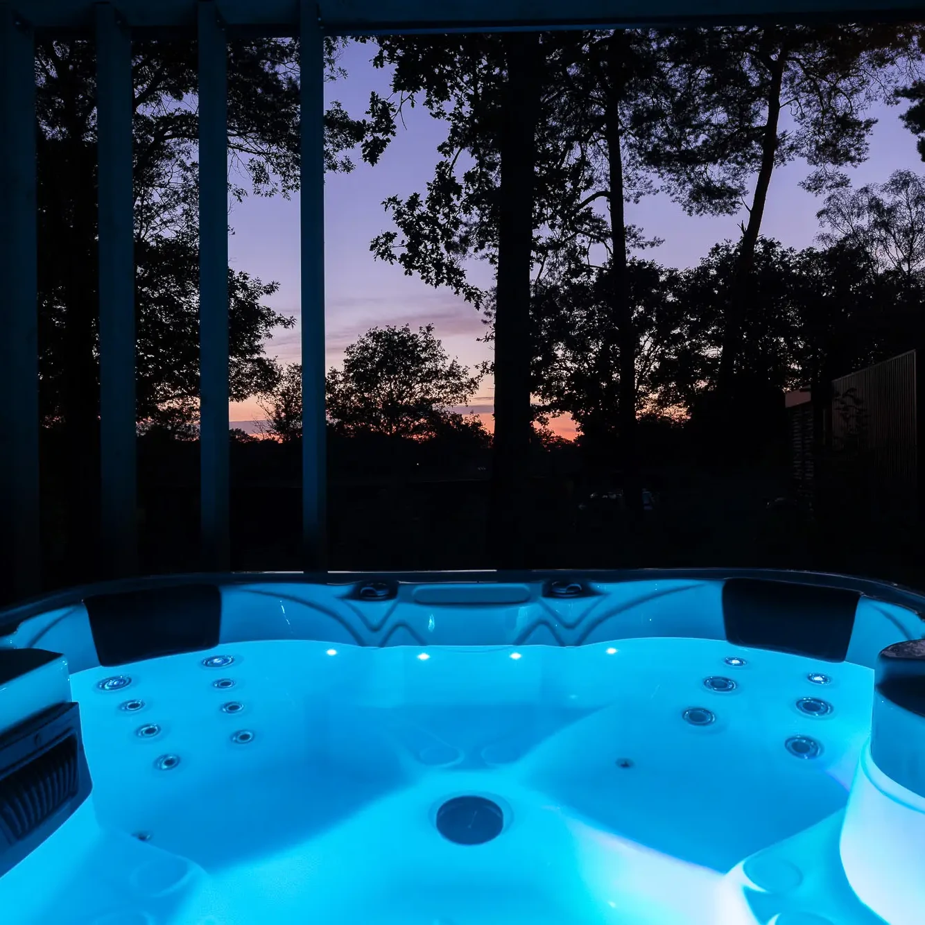 Verlichte jacuzzi met blauw licht bij zonsondergang omringd door bomen bij Zicht op Twente Lodges