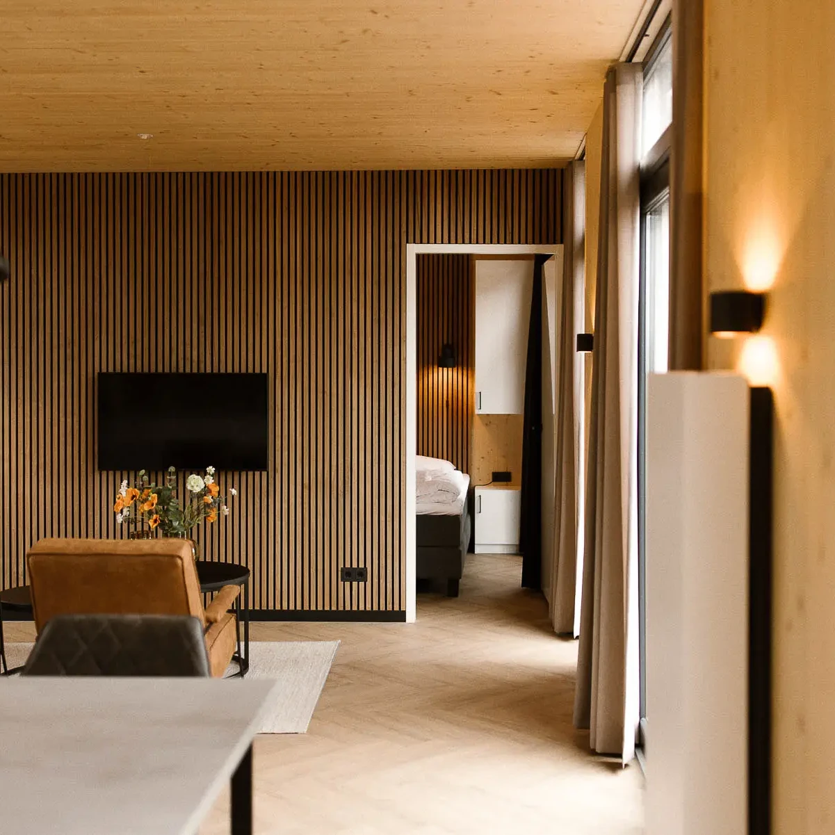 Luxe woonkamer met houten lamellenwand, flatscreen TV en doorkijk naar slaapkamer in Panorama lodge Markelo