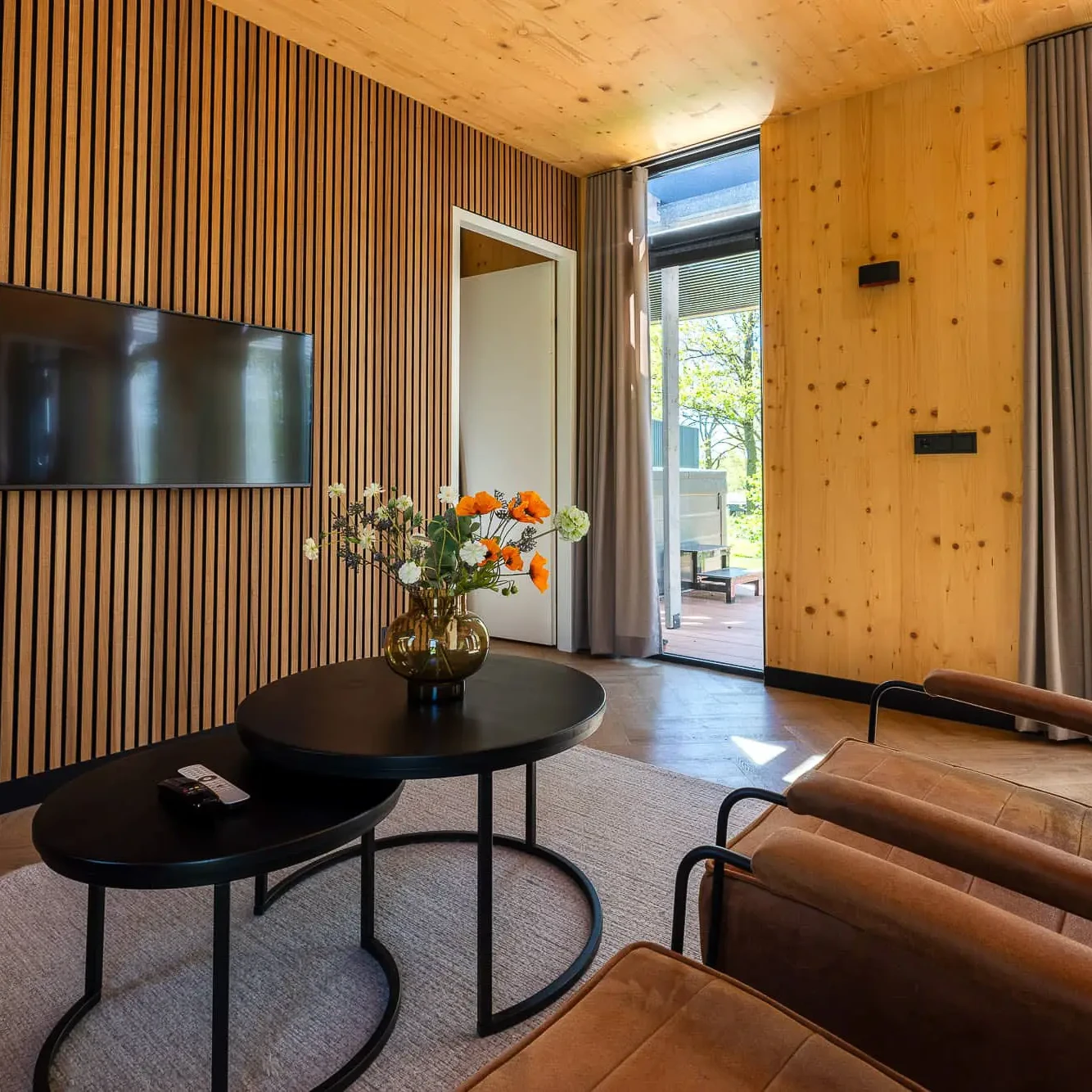 Luxe woonkamer met houten lamellenwand, flatscreen TV en cognackleurige fauteuils in Panorama Lodge Markelo